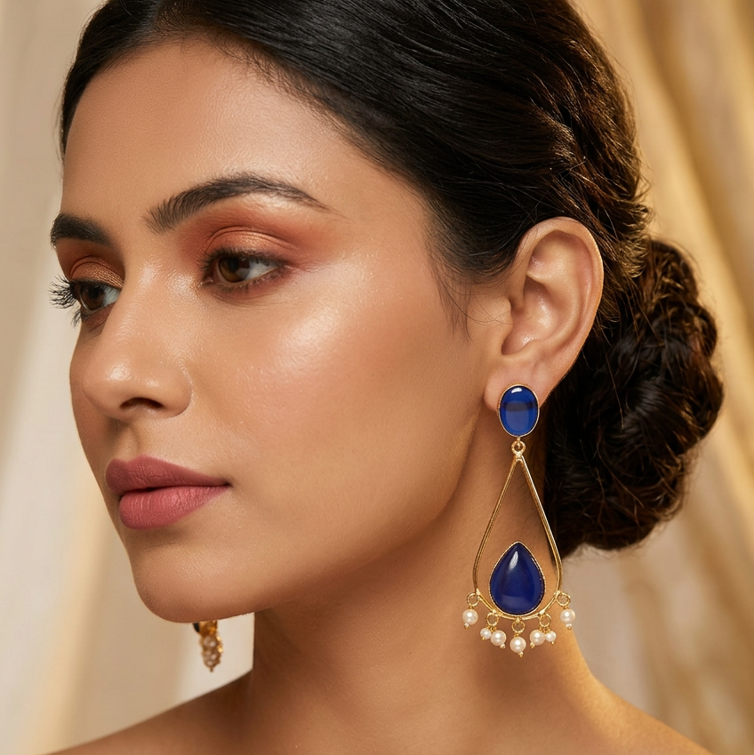 A pair of gold-plated dangle earrings featuring deep blue teardrop-shaped stones and small white pearl accents, made in brass.