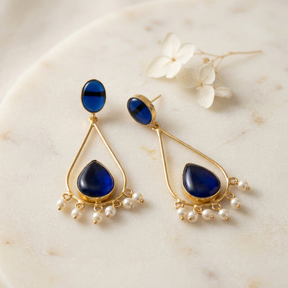 A pair of gold-plated brass dangle earrings featuring deep blue teardrop-shaped stones and small white pearl accents.