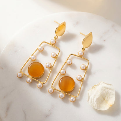 A pair of gold geometric dangle brass earrings with white faux pearls and a large amber-orange circular stone.